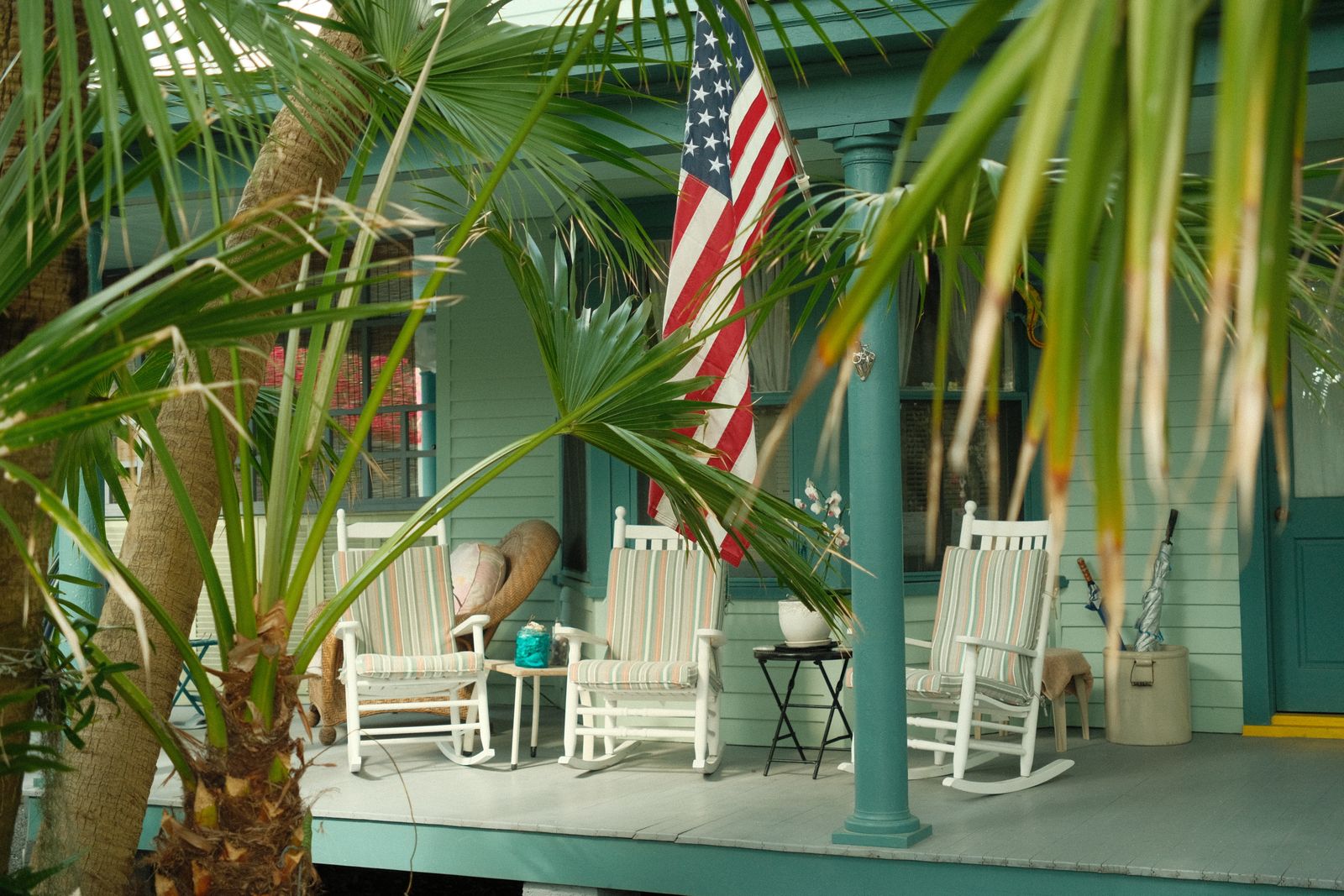 Charming Southern porch with American flag and rocking chairs, hidden behind lush palm trees.